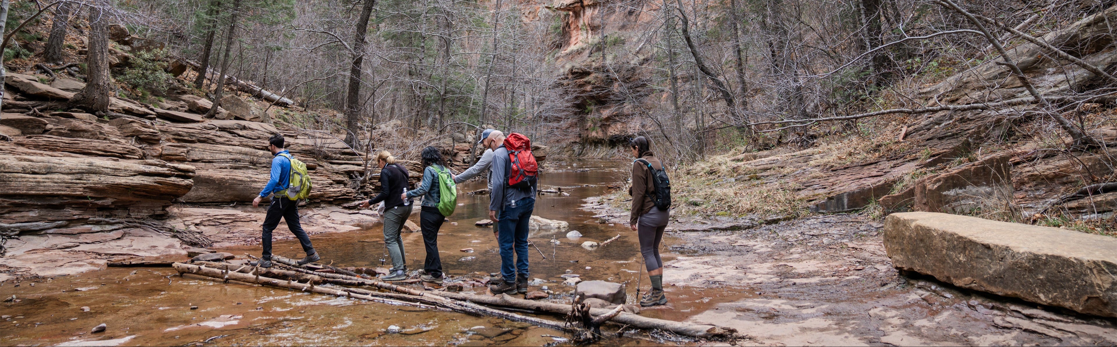 Group of hikers crossing a stream in a forested area with rocky cliffs.